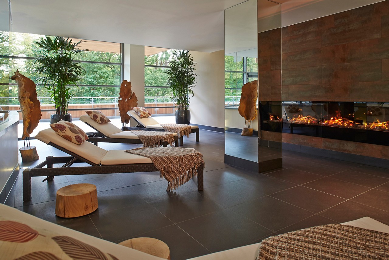Lounge chairs line up facing a long electric fireplace in a serene spa room with large windows, indoor plants, wooden stools, and a mirrored pillar overlooking green trees.